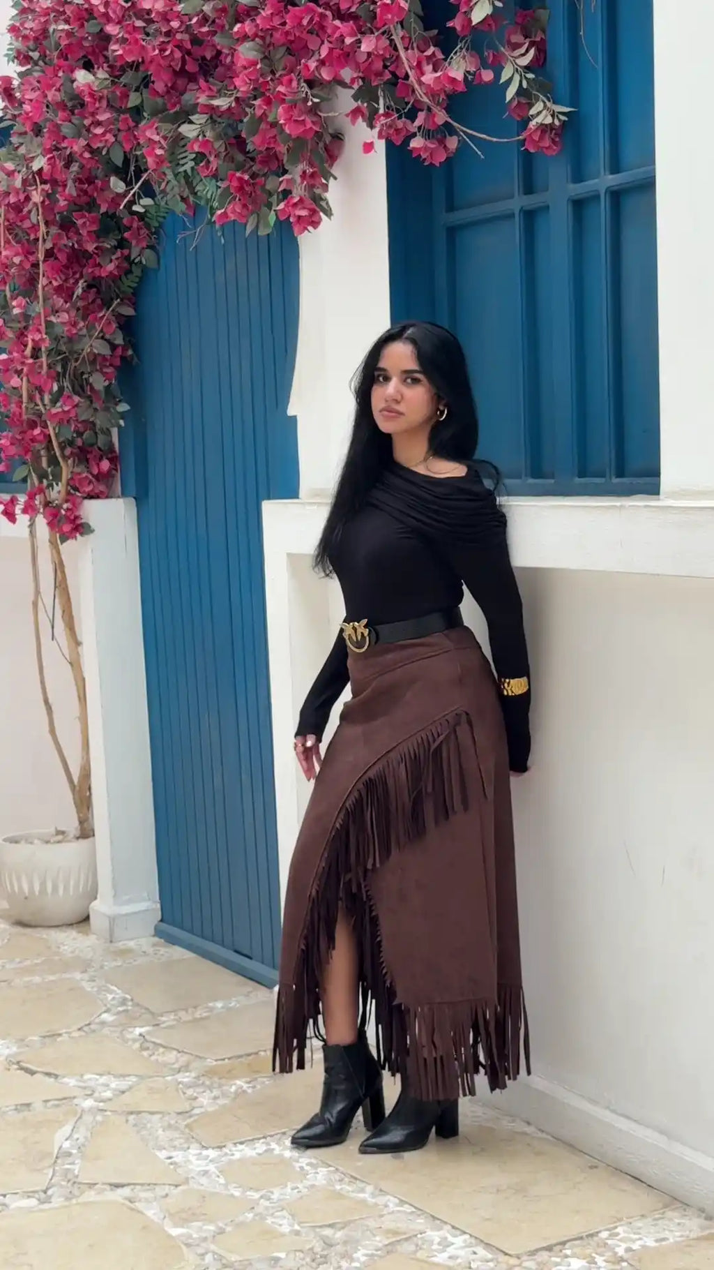 Woman in a black top and brown fringed skirt standing in front of a blue door with pink flowers.