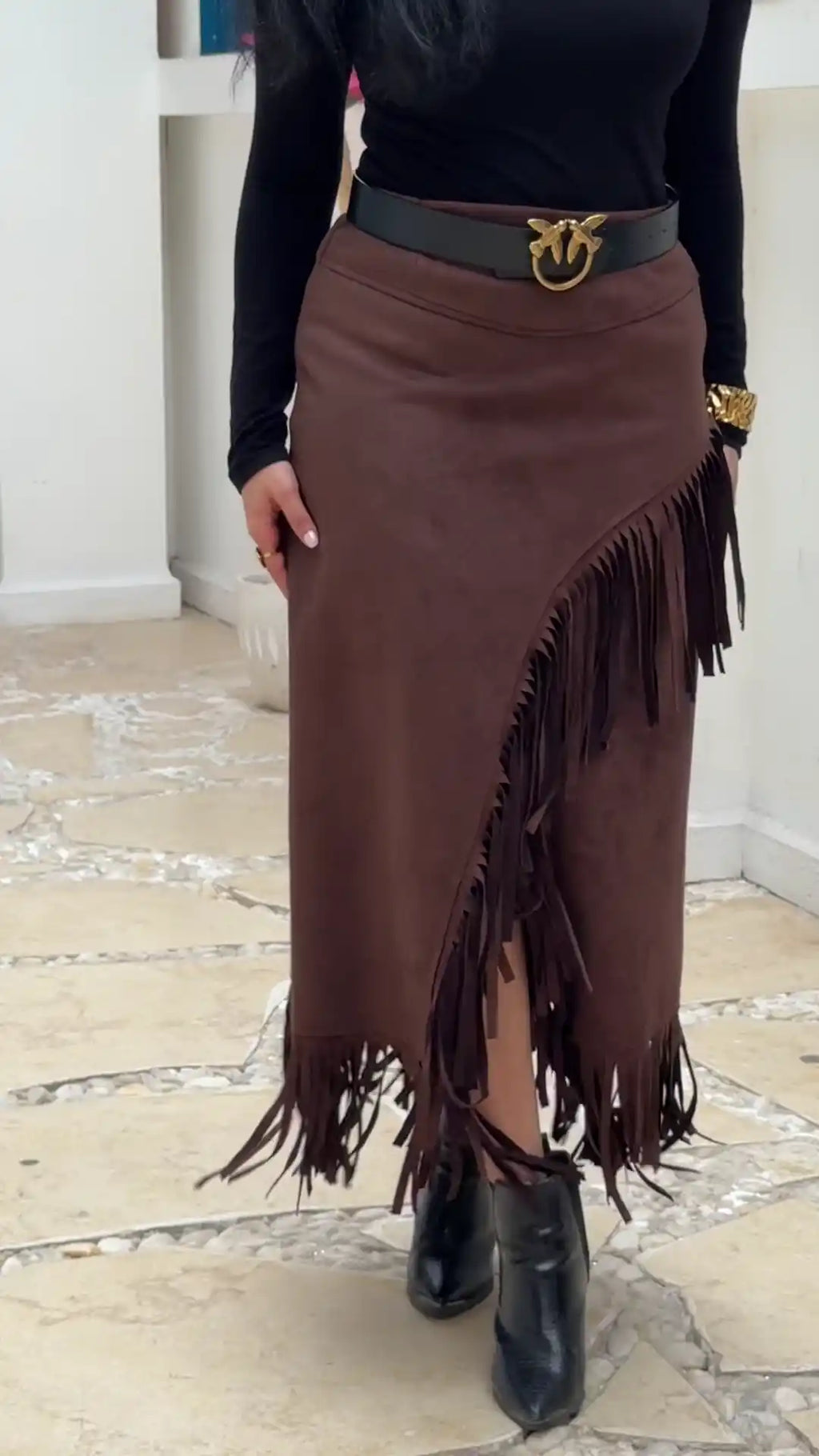 Person wearing a brown fringed skirt with a black belt and black boots on a light wooden floor.
