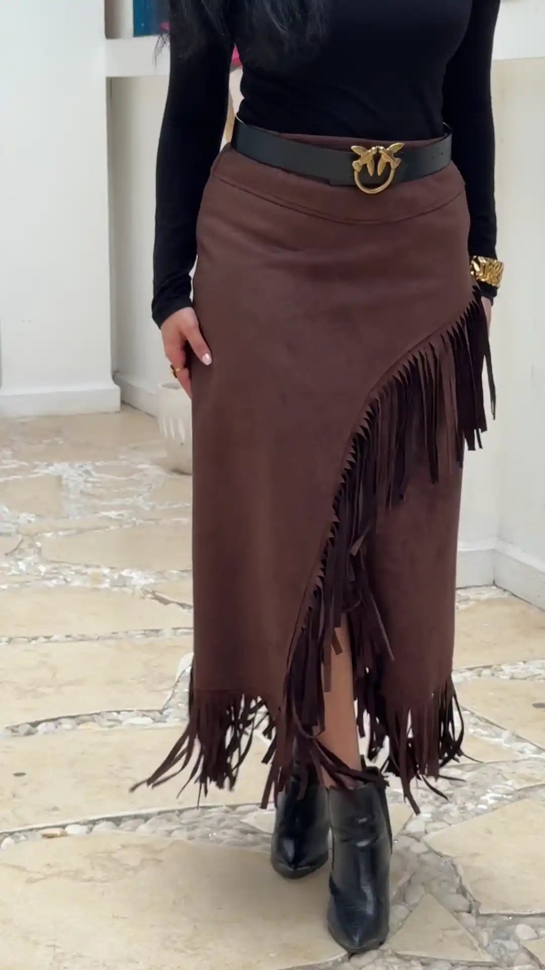 Person wearing a brown fringed skirt with a black belt and black boots on a light wooden floor.