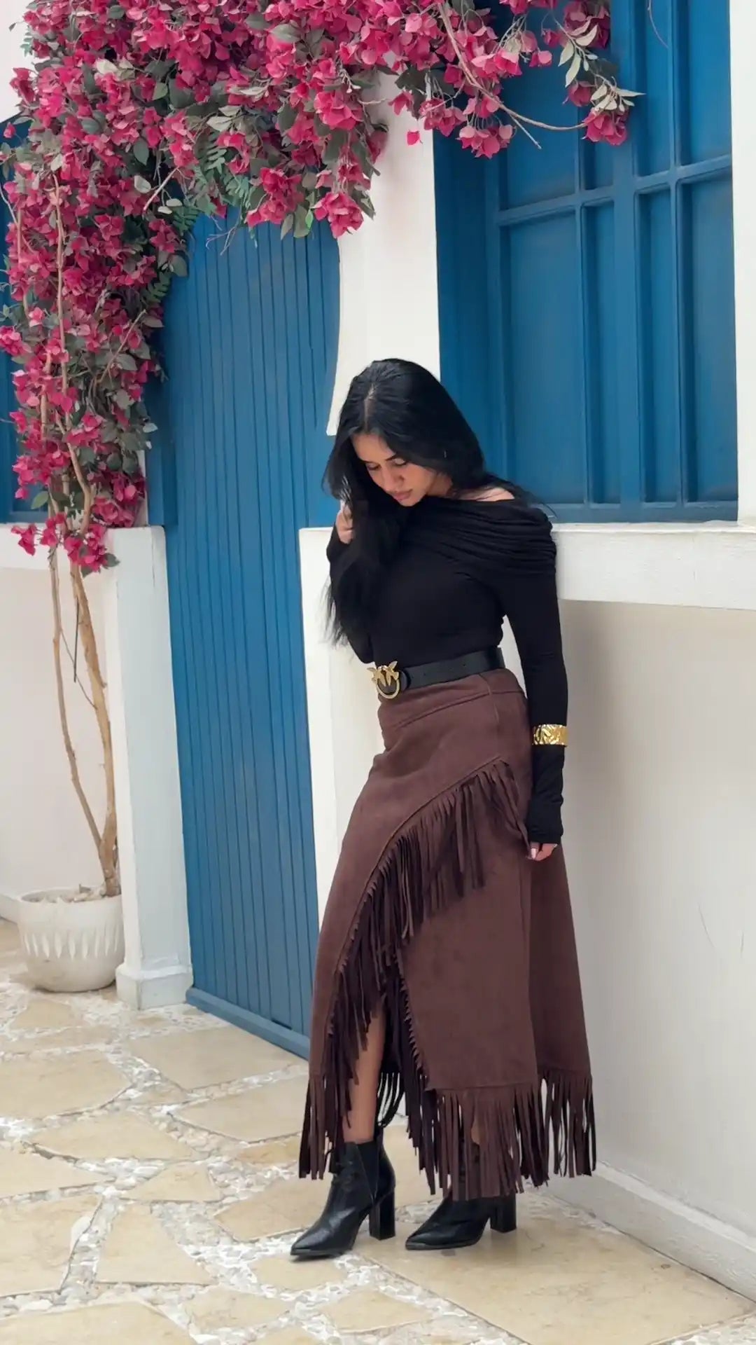 Woman in black top and brown fringed skirt standing in front of a blue door with pink flowers.